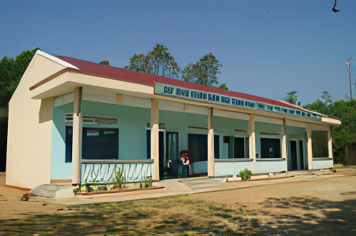 A single-story building with a red roof, light blue and beige walls, multiple windows and doors, and sign text above the entrance, set in a rural environment with trees in the background.