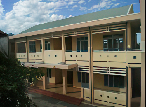 A two-story building with a light yellow exterior and green roof, featuring balconies with railings on both floors and multiple windows under a partly cloudy sky.