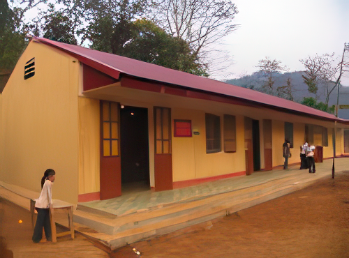 A single-story yellow building with a red roof, featuring several doors and windows. A person walks on a dirt path nearby, and trees are visible in the background.