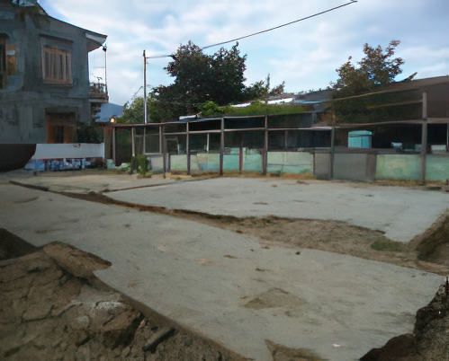 A vacant concrete foundation surrounded by a damaged fence, with residential houses and trees in the background under a cloudy sky.