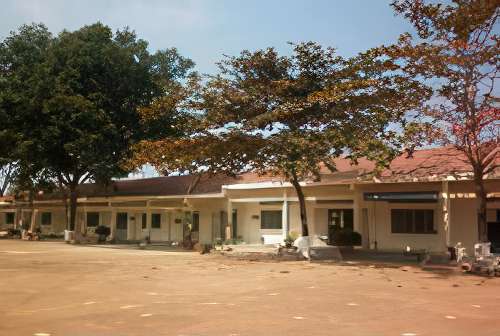 A single-story building with a red-tiled roof, surrounded by trees, and a dirt courtyard in the foreground, under a clear sky.