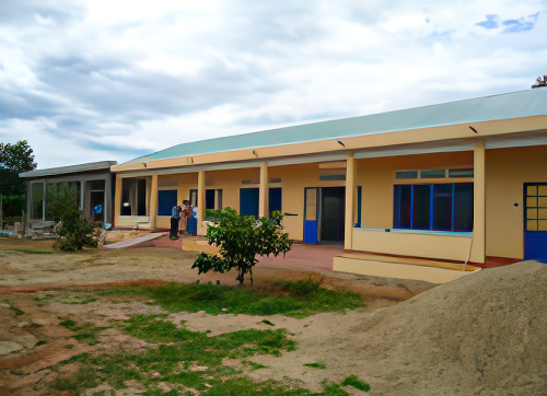 A single-story yellow building with blue doors and windows stands against a cloudy sky. There are a few people near the entrance and some greenery in front of the building.