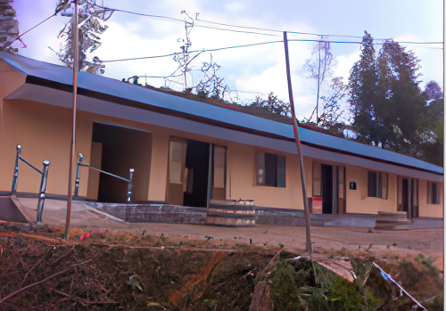 A single-story building with a metal roof, beige walls, open doors, and small concrete steps in front, situated in a rural area with trees in the background.