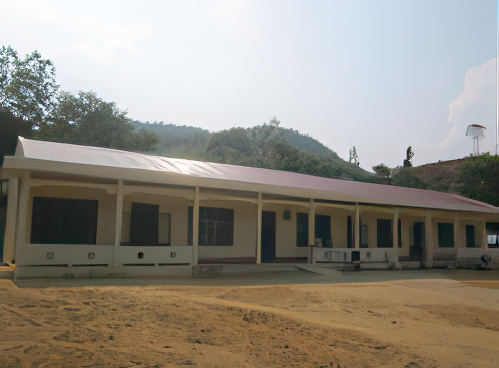 A single-story building with a long front porch and a metal roof stands in a rural, hilly area on a sunny day.