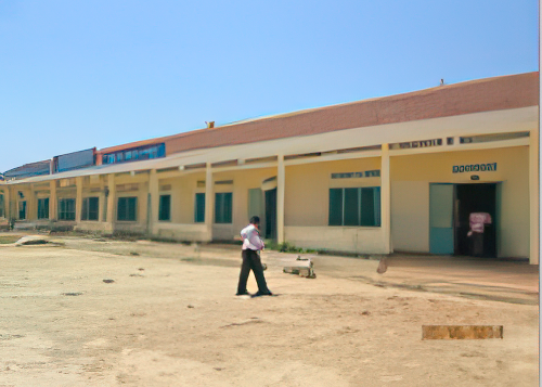 A person stands outside a single-story building with multiple rooms, labeled "Accueil," "Caisse," and "Consultation." The building appears to be part of a medical or administrative facility.