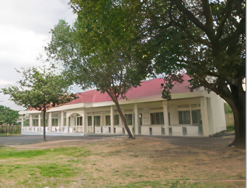 A single-story building with a red roof, white exterior walls, and several large archways at the front. It is surrounded by trees and patches of grass.