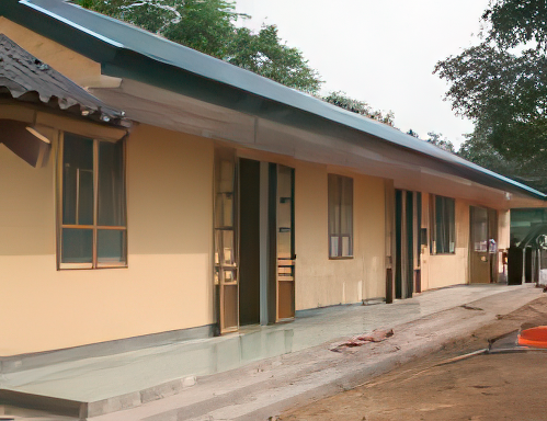 A weathered, single-story building with a slanted roof, wooden pillars, and concrete walls stands in a rural setting with a dirt path and greenery in the background.