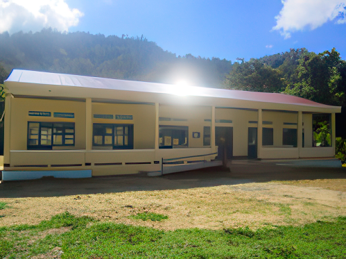 A small, single-story, yellow building with blue windows and an open door, surrounded by trees and a dirt ground.