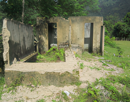 An abandoned, crumbling concrete structure sits amidst overgrown vegetation and grass, with partial walls and remnants of a roof visible against a background of trees and a hillside.