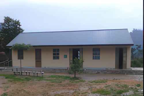 A single-story beige building with a metal roof and multiple doors and windows stands surrounded by a partially grassy area and some trees.