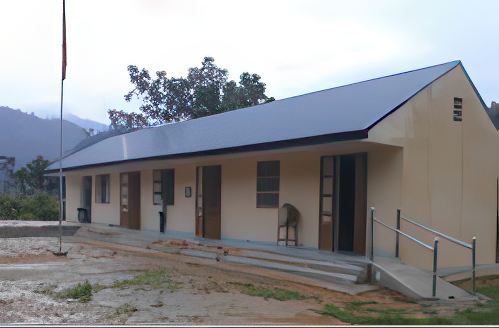 A one-story building with a sloped roof, four doors, and a small ramp leading to the entrance. A flagpole stands in the foreground, and trees and hills are visible in the background.