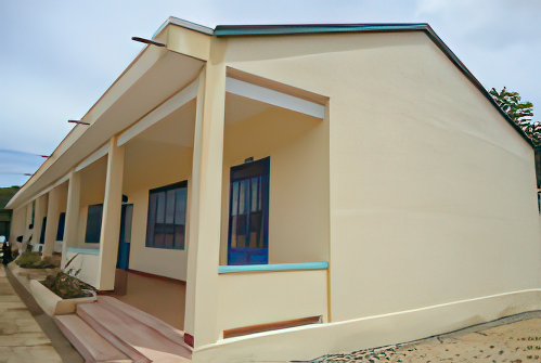 A single-story building with a beige exterior, blue doors, and windows, featuring a covered porch area extending along its front. The sky is partly cloudy in the background.