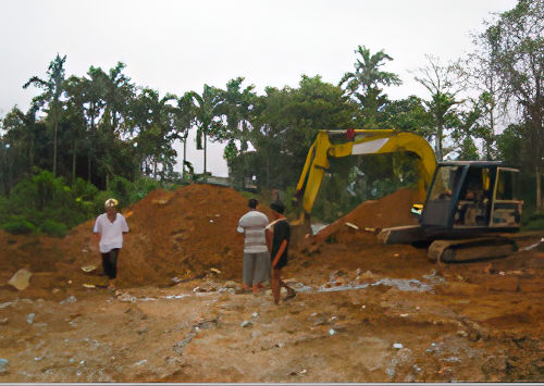 Several people stand near an excavator moving earth at a construction site with trees in the background.