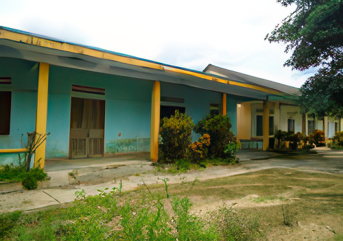 An empty, single-story building with a blue facade and yellow columns stands surrounded by overgrown vegetation. The building appears to be weathered and possibly abandoned.