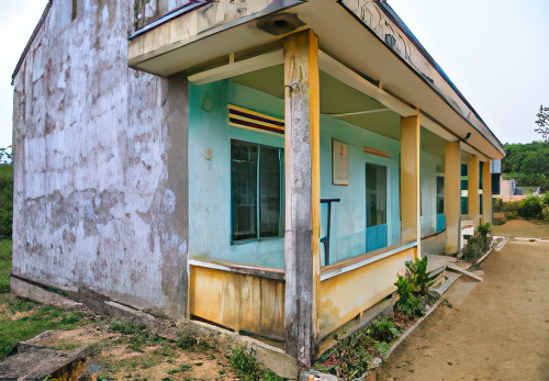 A weathered and partially decayed one-story building with a flat roof, blue walls, yellow-tinted pillars, and a small walkway with overgrown grass on the side.