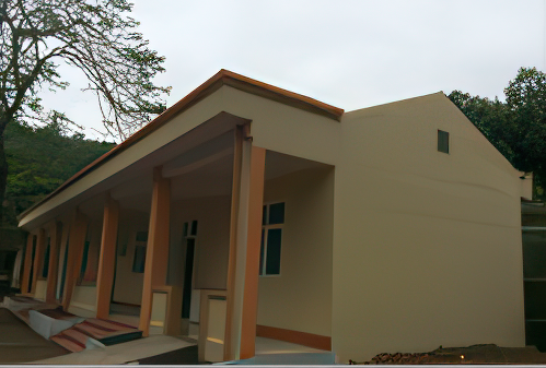 A beige building with orange accents and a slanted roof stands against a backdrop of trees and cloudy sky. The building features several windows and doorways.