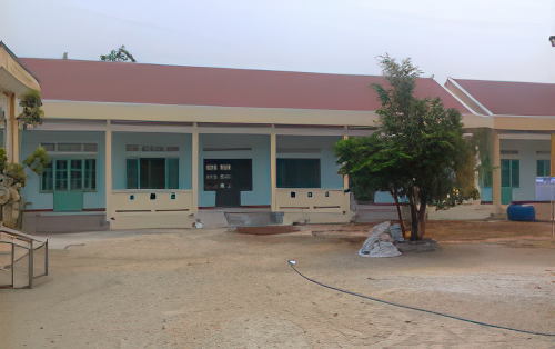 A single-story building with a red roof and green doors and windows stands in a barren courtyard with a few scattered plants and a hose on the ground.