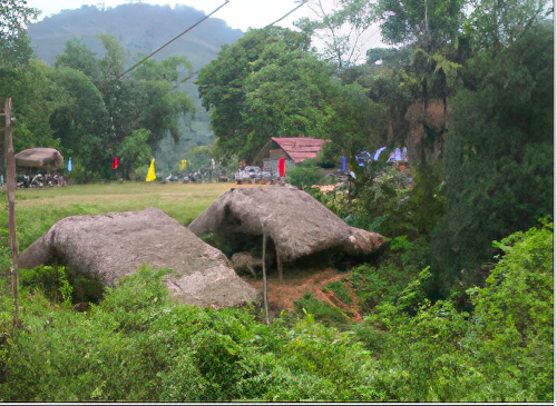 A grassy landscape with two large, flat rocks in the foreground, surrounded by dense greenery, trees, and hills in the background. A small red-roofed structure and scattered items are visible in the distance.