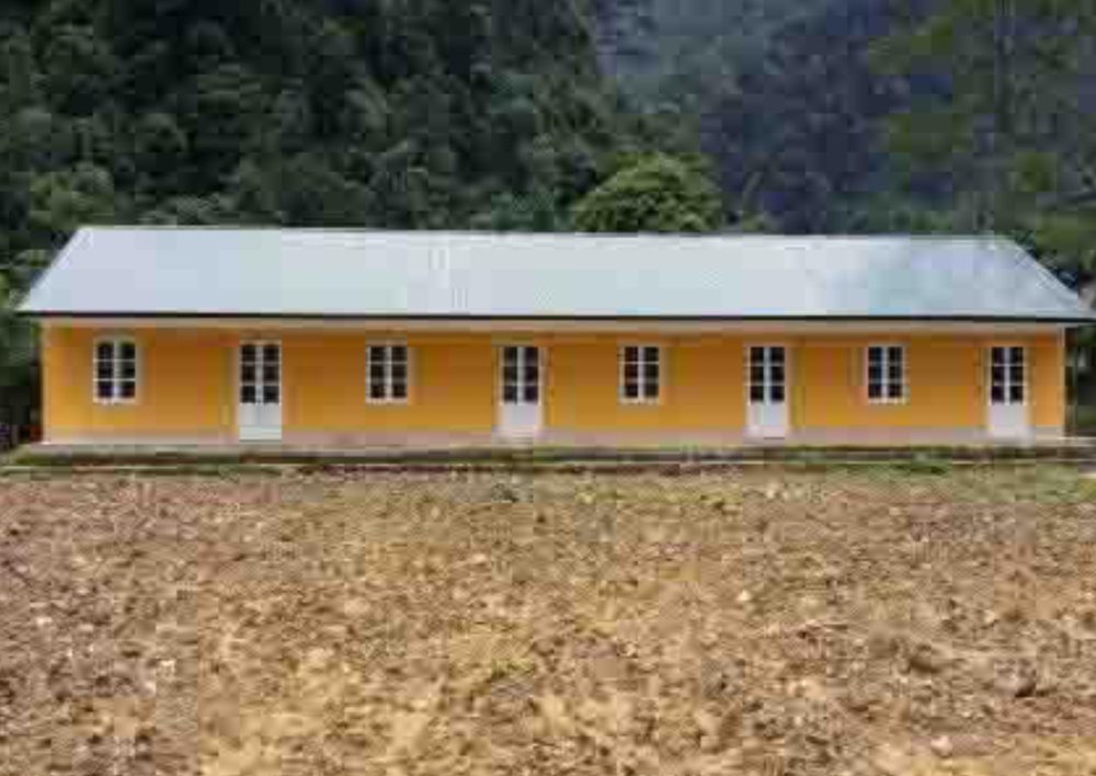 A small, rectangular, yellow building with a white roof, several windows, and doors stands on a cleared patch of land with a forested area in the background.