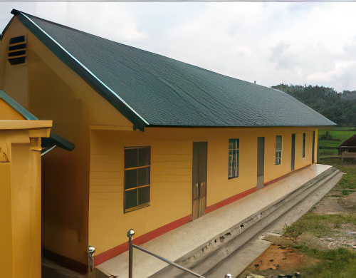 A yellow, single-story building with a green roof is shown. It has multiple doors and windows and is situated in a landscaped area with a concrete pathway leading alongside it.