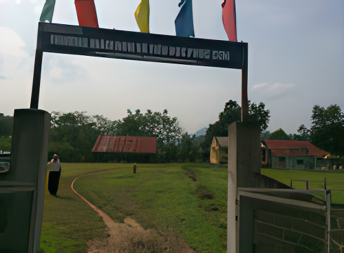 A metal gate with a sign in a non-Latin script opens to a grassy path leading to buildings with red roofs surrounded by trees. There are colorful flags on top of the gate.