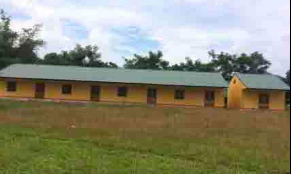 A single-story building with a green roof, yellow walls, and multiple evenly spaced doors and windows, situated in a grassy area with trees in the background.