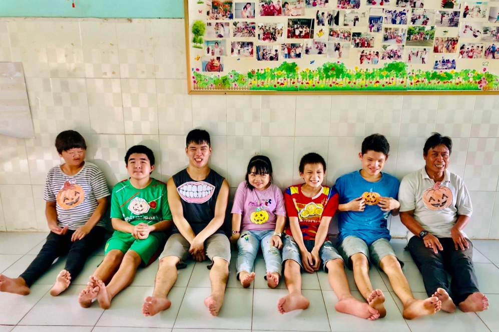 Six smiling children and one man sit on the floor in a tiled room, their legs stretched out. They wear bright clothes with paper cutouts pinned to their shirts. A photo collage is on the wall behind them, showcasing moments from their journey with rehabilitation services.