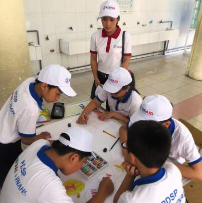 Children wearing USAID caps and shirts collaborate on a drawing at a table. An adult in a similar outfit supervises. The setting appears to be an indoor area with white tiled walls.