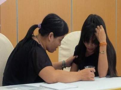 Two women sitting at a table indoors, one woman comforting the other as she holds her head with one hand and a drink in the other.