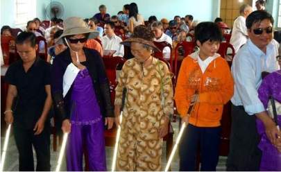 A group of visually impaired individuals using white canes are standing in a line inside a room filled with seated people in the background. Some wear sunglasses and hats.