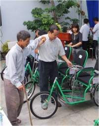 Two men inspecting green tricycles for the elderly and disabled at an outdoor event. One man is holding a cane, and there are several people in the background.