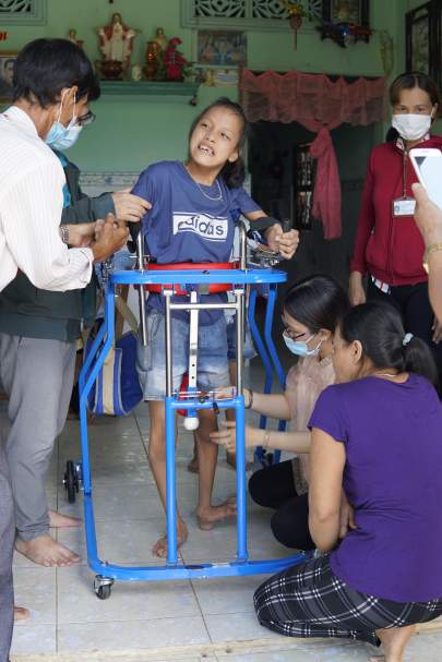 A child in a blue t-shirt uses a mobility aid with the support of several adults in a room. The adults, skilled in various therapies, are assisting and adjusting the child's position, highlighting the importance of disability rights.