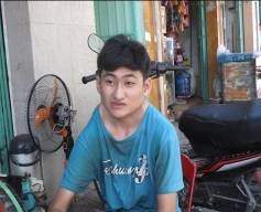 A young person wearing a blue shirt sits near a motorcycle and a fan, outdoors beside a building.