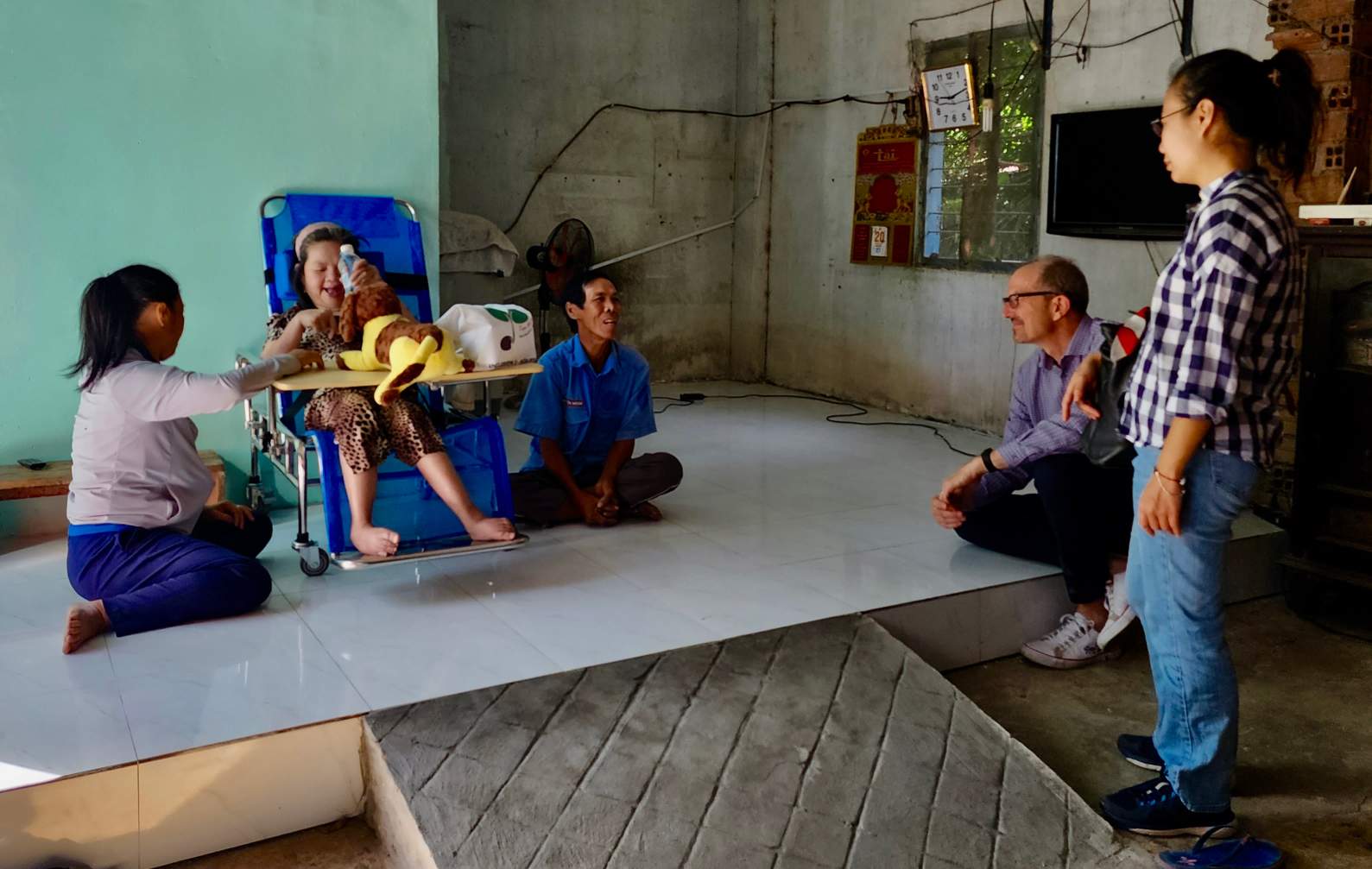 A group of people interact with a smiling child on a wheelchair, benefiting from rehabilitation services in a modest room with concrete walls and a small portable TV.