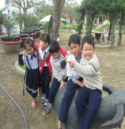 Five children in school uniforms are playing on a statue of an animal in an outdoor setting. There are trees, potted plants, and a park bench in the background.