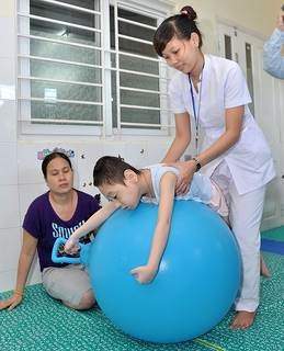 A physical therapist guides a child on a large exercise ball, with another woman sitting nearby in a room with a window and tiled walls.