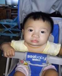 A baby sits in a blue stroller labeled "seebaby." The child has short black hair and is wearing a yellow shirt. There is furniture and other objects visible in the background.