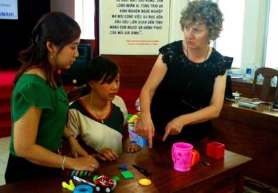 Three women are engaged in a discussion while examining various small objects on a table in an indoor setting. One woman is pointing at something on the table.