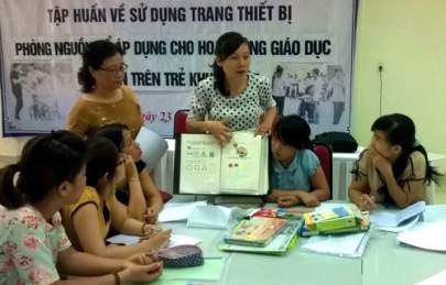 A woman holds up a book and explains its contents to a group of seated people in a classroom setting. A whiteboard and a banner with Vietnamese text are in the background.