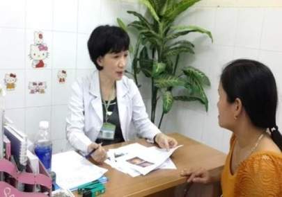 A doctor in a white coat is seated at a desk, talking to a woman in an orange top. They are in a medical office with some decorative stickers and a potted plant in the background.