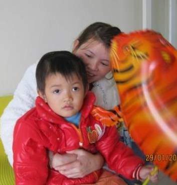 A young child in a red jacket holds a tiger balloon while being embraced by a woman. Both are sitting on a couch. 