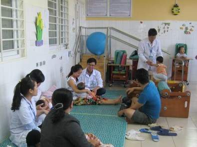 A group of adults and children are in a room with white walls. Some adults are seated on mats, holding children, while others assist and interact with the children. Medical personnel in white coats are present.