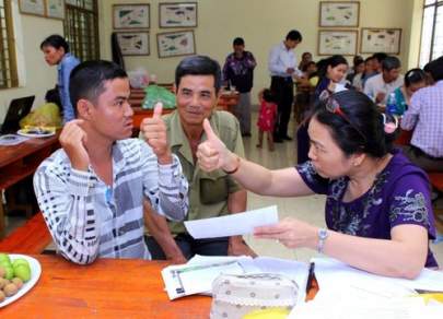 A seated woman shows a thumbs-up to a seated man in a striped shirt, who also gives a thumbs-up. Another man in a green shirt observes. Several people work at tables in the background.