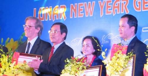 Four people stand in a row on stage holding awards, with a backdrop that reads "Lunar New Year." They are dressed in formal attire and there are yellow flowers in the foreground.