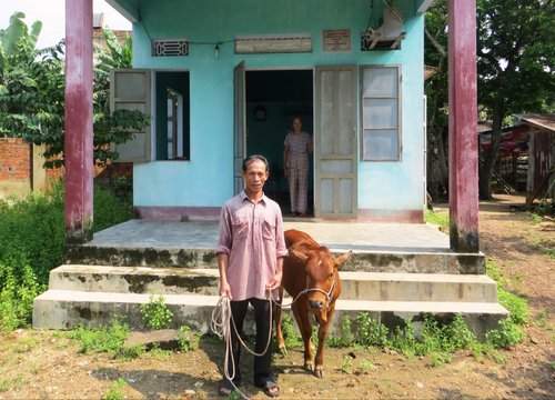 A man stands in front of a light blue house holding a rope tied to a brown cow. A woman stands in the doorway behind him.