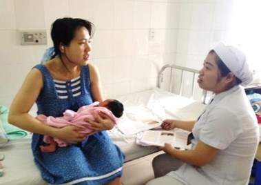 A woman holding a baby sits on a hospital bed, speaking with a nurse who is taking notes.