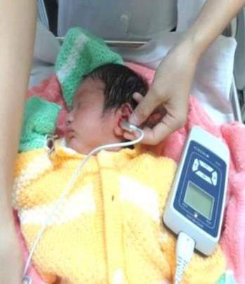 A newborn wrapped in a yellow and pink blanket is undergoing a hearing test with a device held by a healthcare professional.